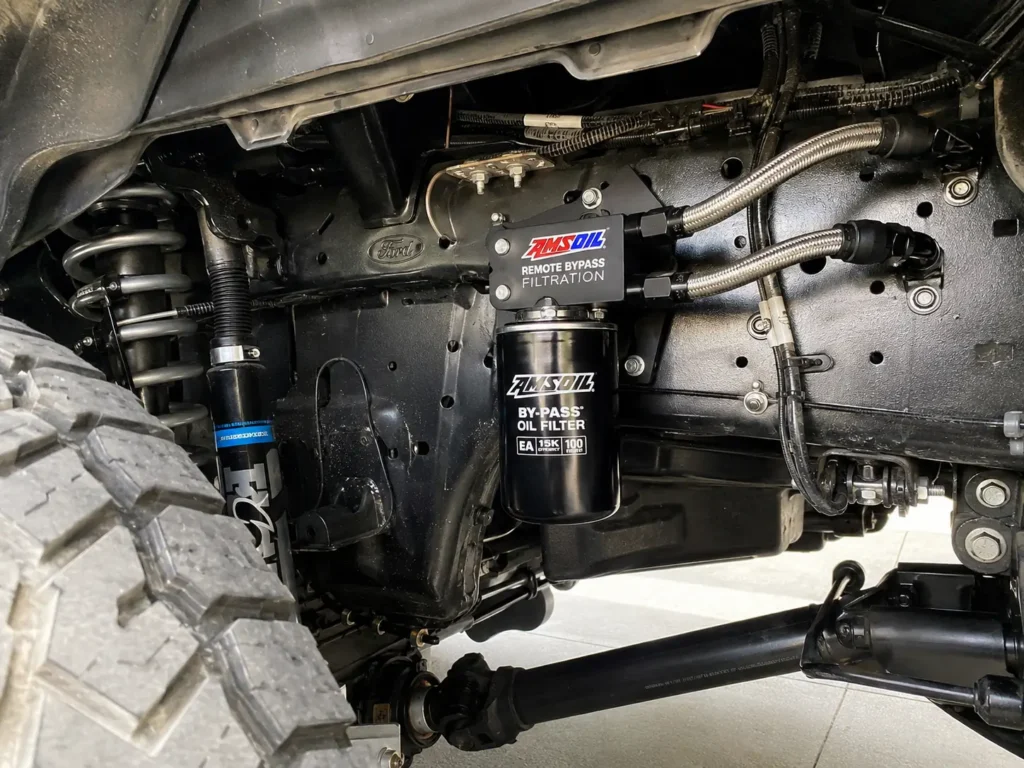 AMSOIL remote bypass oil filter system mounted on a Ford Super Duty frame rail with braided stainless lines behind the front driver-side wheel.