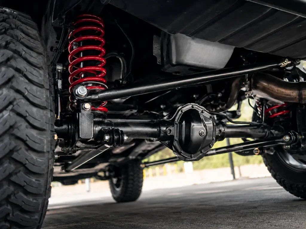 Low-angle undercarriage view of a heavy-duty pickup truck showing the rear axle, track bar, and heavy-duty red rear coil spring suspension in sharp focus.
