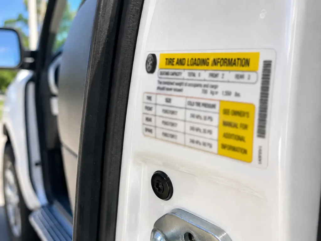 Close-up of an open truck door jamb showing a yellow Tire and Loading Information sticker in bright daylight with shallow depth of field.